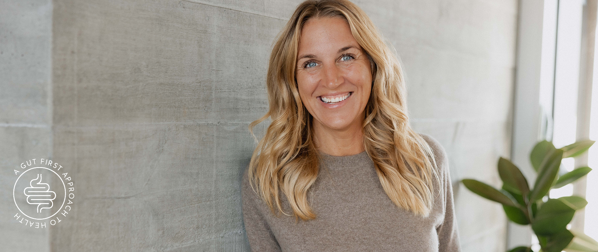 Woman with blonde hair smiling in a room with a plant and light-colored wall.
